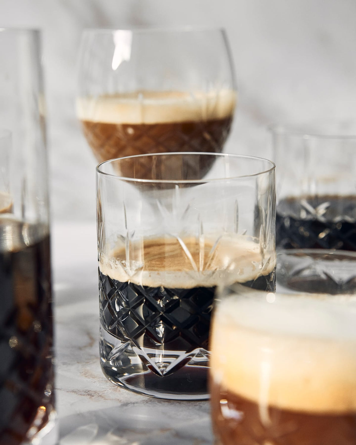 Facetslebet whiskyglas med mørk drink og isterning, omgivet af glas med gyldne drikke