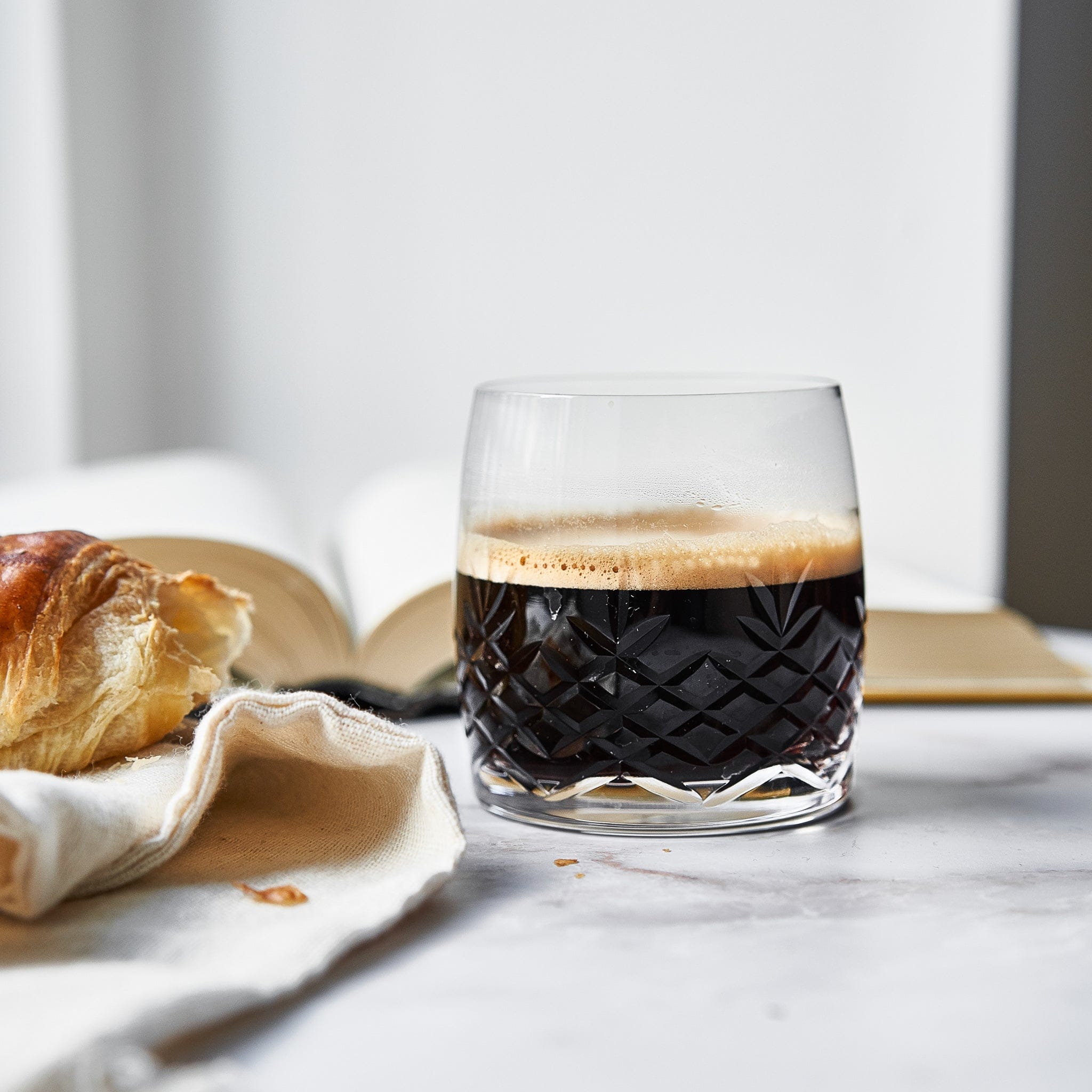 Klar krystalglas med facetslebet diamantmønster fyldt med kaffe ved bog og croissant fra Frederik Bagger