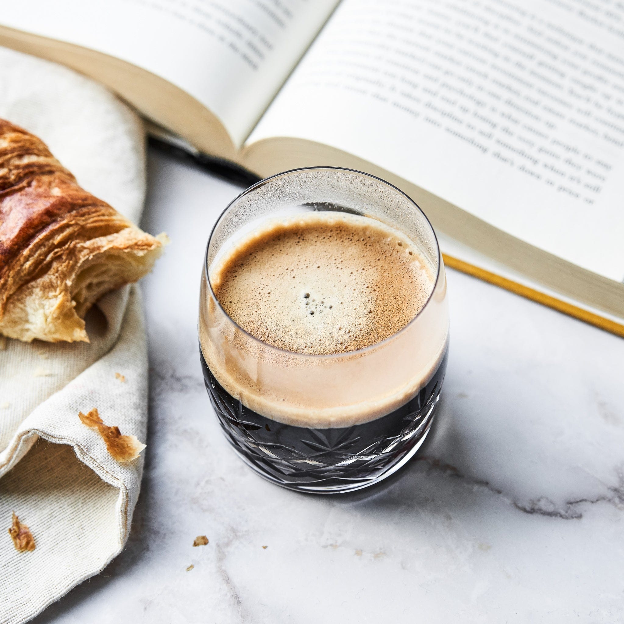 Klar krystalglas med riflet og facetslebet mønster fyldt med kaffe ved croissant og åben bog fra Frederik Bagger