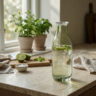 Klar glasflaske med rund silhuet og låg, anvendt til servering med lime og urter på bord fra Frederik Bagger