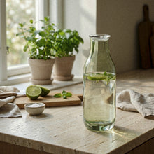 Klar glasflaske med rund silhuet og låg, anvendt til servering med lime og urter på bord fra Frederik Bagger