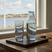 Klar glasflaske med rund silhuet og låg samt to blå, cylinderformede glas på træbakke til servering fra Frederik Bagger